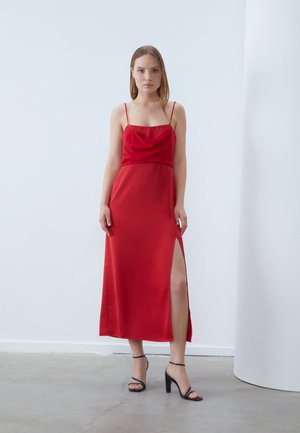 Rochie de seară din satin roșu, cu decolteu tip cowl, bretele subțiri și o fanta înaltă pe coapsă. Sandalele negre cu bretele înalte completează ținuta.