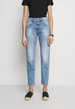 Person wearing light blue faded high-waist jeans, black short-sleeve top, black espadrille shoes, and black wristband, standing on gray floor.