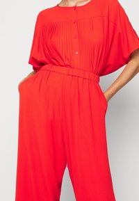 Mono rojo con mangas cortas, sección superior con plisados, cierre de botones en la parte delantera y una cintura elástica. Cuenta con bolsillos laterales y piernas de corte suelto.