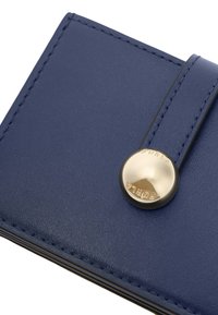 Angolo di un portafoglio in pelle blu con una cinghia cucita e un bottone rotondo dorato inciso con il logo "FURLA".
