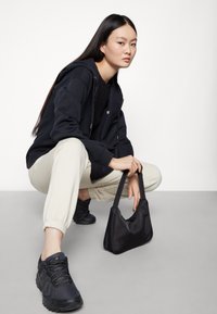 Schwarze Nylon-Handtasche mit geschwungener Form, getragen mit einem schwarzen Hoodie mit Reißverschluss und hellbeigen, schmal zulaufenden Jogginghosen, kombiniert mit schwarzen Sneakers.