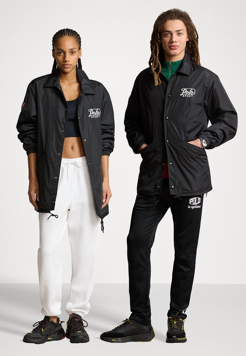 Schwarze Windjacken mit vorderen Knopfverschlüssen und Logo-Akzenten. Die Modelle tragen weiße und schwarze Jogginghosen mit kontrastierenden Streifen und Sneakers.