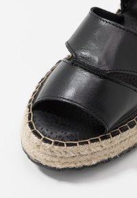 Černé kožené sandály s otevřenou špičkou, se splétanou jutu platformou a viditelným švem na bílém pozadí.