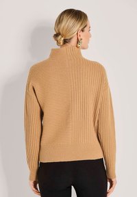 Frau mit blonden Haaren in einem tiefen Dutt, die einen tanfarbenen, gerippten Pullover mit gemischten Strickmustern, schwarze Hosen und auffällige goldene Ohrringe trägt, mit dem Rücken zur Kamera.