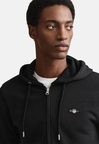 Felpa nera con zip e cappuccio con coulisse, con logo ricamato sul petto e tessuto liscio e strutturato.