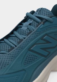 Κοντινό πλάνο ενός αθλητικού παπουτσιού σε τιρκουάζ μπλε με δίχτυ, με κορδόνια στο ίδιο χρώμα και μεγάλο ανάγλυφο λογότυπο "N" στο πλάι πάνω από λευκή σόλα.