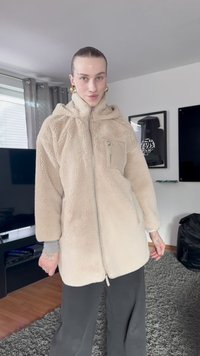 Beige fleece jacket with a hood, front zipper, and a small chest pocket. Textured, loose fit with long sleeves, worn over dark pants.