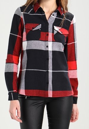 Femme portant une chemise à carreaux rouge, noire et blanche à boutons avec poches avant, associée à un pantalon en cuir noir.