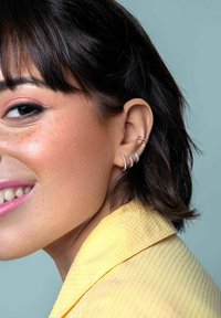 Trois boucles d'oreilles argentées de différentes formes sur l'oreille d'un modèle, sur fond bleu doux, avec un vêtement texturé jaune.