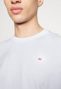 T-shirt bianco in cotone con scollatura rotonda e un piccolo logo rosso e bianco sul lato sinistro del petto. Texture liscia, design minimale.