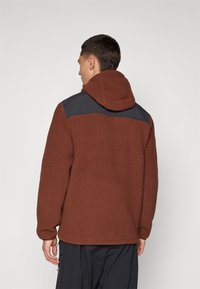 Veste en polaire marron avec une texture douce, dotée d'un empiècement noir aux épaules et d'une capuche. Les poignets sont légèrement ajustés, offrant du confort.