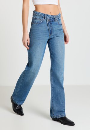 Donna che indossa jeans blu a vita alta con gamba dritta e chiusura asimmetrica unica con bottoni, abbinati a stivali neri con punta affilata.