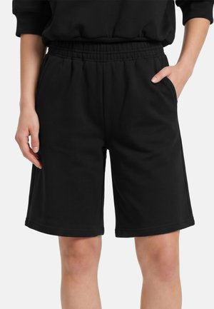 Czarne, sięgające do kolan szorty z elastycznym pasem i bocznymi kieszeniami, noszone przez osobę z jedną ręką w kieszeni, a drugą luźno ułożoną.