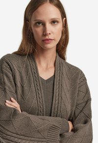 Grau gestrickte Strickjacke mit einem strukturierten Zickzackmuster, ausgestattet mit einem Schalkragen und langen Ärmeln über einem passenden V-Ausschnitt-Top.