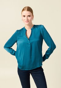 Blusa lunga teal con maniche lunghe e scollatura a V, con dettagli a bottone. Realizzata in tessuto liscio, presenta una vestibilità rilassata con un orlo leggermente curvo.