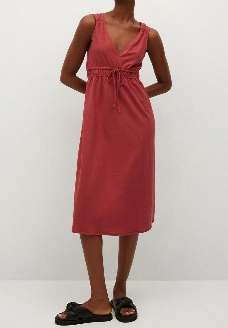 Femme portant une robe sans manches, longueur au genou, couleur rouille avec une taille à cordon et des sandales noires à enfiler, debout devant un fond uni.