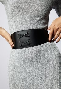 Ceinture large en simili cuir noir avec détail à lacets et œillets en métal, portée sur une robe grise texturée. Fini lisse avec un design épuré.