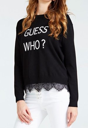 Vrouw draagt een zwarte trui met de tekst "GUESS WHO?" en kanten zoom, gecombineerd met witte hoge taillebroek met vetersluiting.