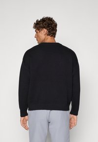 Svart strikket genser med senkede skuldre, ribbestrikkede mansjetter og kant, samt en avslappet passform, vist fra baksiden på en modell.