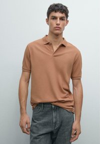 Young man standing against plain wall wearing a brown polo shirt and faded black jeans, looking straight ahead with a neutral expression.