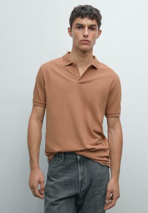 Junger Mann steht gegen eine schlichte Wand, trägt ein braunes Polohemd und ausgebleichte schwarze Jeans, schaut geradeaus mit einem neutralen Gesichtsausdruck.