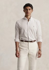 Hvit, stripete skjorte med knapper laget av bomull, kombinert med lyse bukser. Merkbar brun lærbelte og klokke. Minimalistisk design.