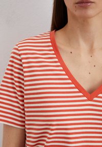 Rot- und cremefarbener gestreifter T-Shirt mit V-Ausschnitt. Weicher Stoff und kurze Ärmel. Auffällige glatte Textur und gleichmäßiges Streifenmuster.