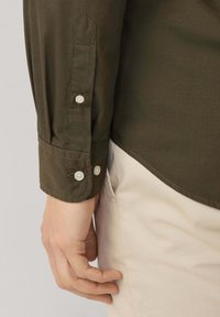 Camisa verde oliva de manga larga, con puños de botón y dobladillo curvado. La tela parece suave con una textura robusta. Los botones son blancos.