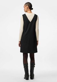 Černé šaty bez rukávů s výstřihem do V na zádech, doplněné krémovým topem s dlouhými rukávy a černými kozačkami po kolena se sponou, tvořící vrstvený vzhled.