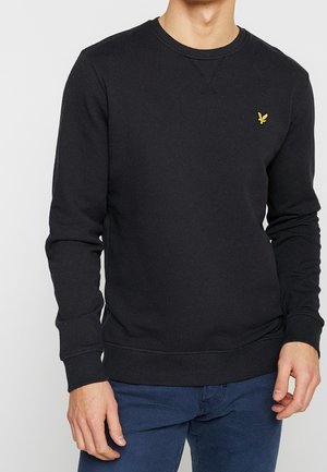Homme portant un sweat-shirt noir à col rond avec un petit logo d’oiseau jaune sur la poitrine et un pantalon bleu foncé.