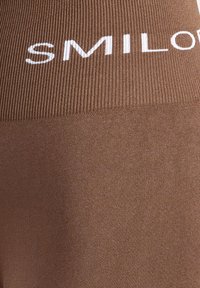 Leggings marrones con cintura acanalada que presenta el texto blanco "SMILOR." El material parece suave con una textura consistente y un brillo sutil.