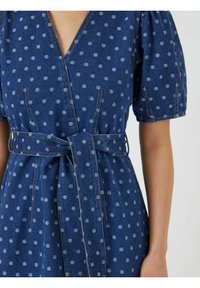 Denim mekko syvällä V-kaula-aukolla, puffihihnoilla ja kapealla vyötäröllä. Ominaisuuksiin kuuluu sininen kangas, jossa on vaaleansiniset pilkut ja näkyvät saumat.