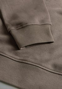 Světlý hnědý sweatshirtový materiál s ribovanými manžetami a lemem. Má hladkou strukturu a jemné detaily šití.