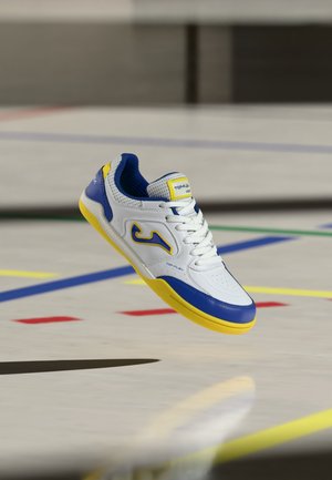 Chaussure de sport blanche, bleue et jaune flottant au-dessus d’un sol de gym avec des lignes de terrain colorées en arrière-plan.