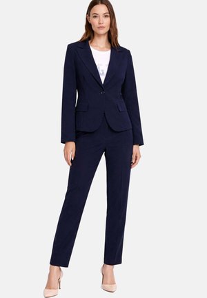 Donna in piedi che indossa un blazer blu scuro aderente e pantaloni dritti abbinati, con una camicia bianca e tacchi beige, su uno sfondo neutro.