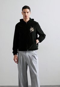 Casaco de veludo preto com fecho éclair e capuz, com um design bordado de cobra dourada no peito, usado sobre calças plissadas cinza claro.