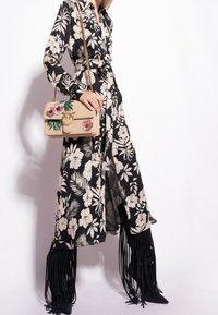 Kobieta w długiej czarnej sukience w kwiaty, trzymająca beżową torebkę ozdobioną kwiatami, w wysokich czarnych botkach z frędzlami, na białym tle.