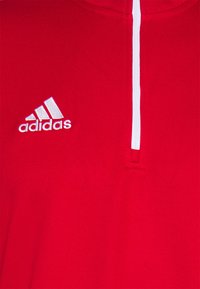 Punainen villapaita, jossa on puoliveto-aukko, valmistettu teksturoidusta kankaasta. Vasemmassa rinnassa on valkoinen Adidas-logo.