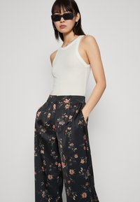 Top in tessuto aderente bianco con scollatura rotonda; abbinato a pantaloni ampi in satin nero con motivo floreale, caratterizzati da pieghe e tasche laterali. Occhiali da sole neri.
