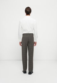 Mann, der mit dem Rücken zur Kamera steht, gekleidet in einem weißen Langarmshirt, grauen karierten Hosen und schwarzen Schuhen vor einem schlichten weißen Hintergrund.