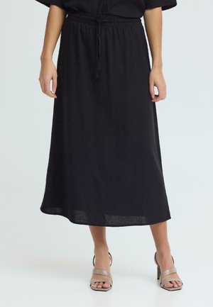 Kvinne iført svart midiskjørt med elastisk midjebånd og snøring, kombinert med beige åpne blokkhælte sandaler på hvit bakgrunn.