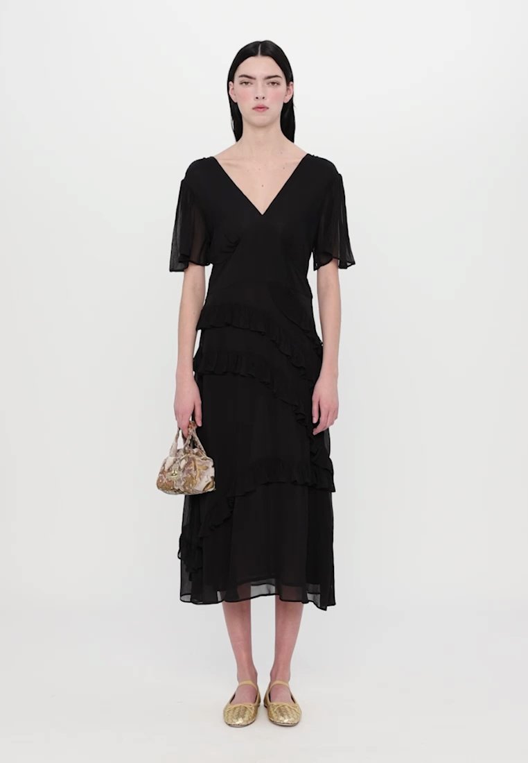 Femme se tenant debout, portant une robe midi noire à volants, tenant un sac à main beige à motifs et portant des chaussures plates dorées.