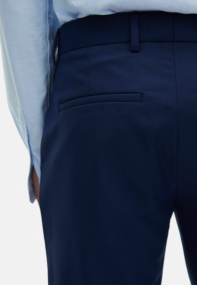 Dunkelblaue Hosen aus strukturiertem Stoff mit einer verdeckten Tasche, kombiniert mit einem hellblauen Hemd. Der taillierte Schnitt zeigt eine schräg verlaufende Taille.