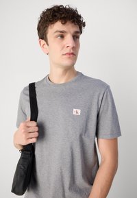 Jaunas vyras garbanotais plaukais, dėvintis pilką Calvin Klein marškinėlį ir laikantis juodos peties krepšio dirželį, žiūri į šoną.