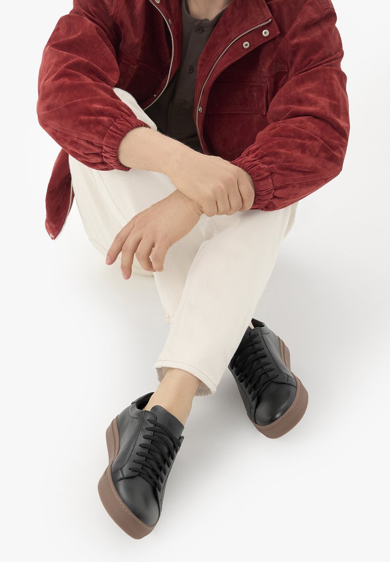 Personne assise les mains jointes, portant une veste rouge, un pantalon beige et des baskets noires avec des semelles marron sur fond blanc.