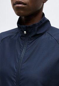 Κοντινό πλάνο ενός ατόμου που φοράει μπλε ζακέτα με φερμουάρ και όρθιο γιακά, μερικώς ανοιχτή, που δείχνει το κάτω μέρος του προσώπου και τον λαιμό.