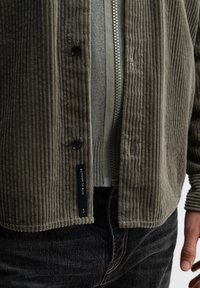 Olivenfarbene Cord-Jacke mit vertikalen Rippungen, Knopfverschluss und Etikett. Die innere Schicht besteht aus einem reißverschlussartigen, hellgrünen Material.