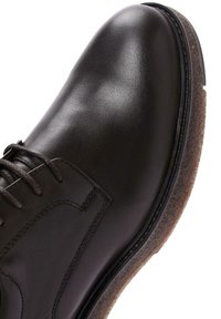 Bottine en cuir marron avec une finition lisse, bout rond et accents cousus. Présente des lacets et une semelle texturée marron.