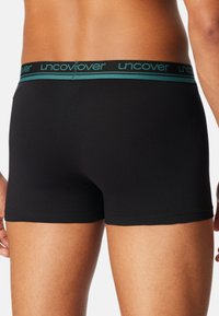 Svarta boxershorts i glansigt tyg, med en elastisk midjeresår accentuerad i teal och "uncover" märkning. Tätt sittande design.