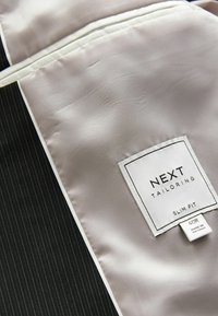 Czarna tkanina w paski z gładką beżową satynową podszewką. Biała metka z napisem "NEXT TAILORING SLIM FIT", wskazująca rozmiar i pochodzenie.